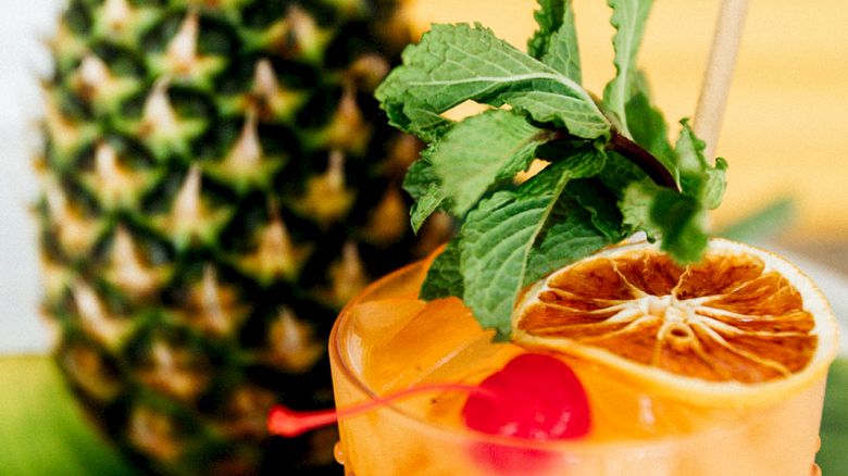 A tropical cocktail garnished with mint, orange slice, and cherry sits next to a pineapple on a banana leaf.