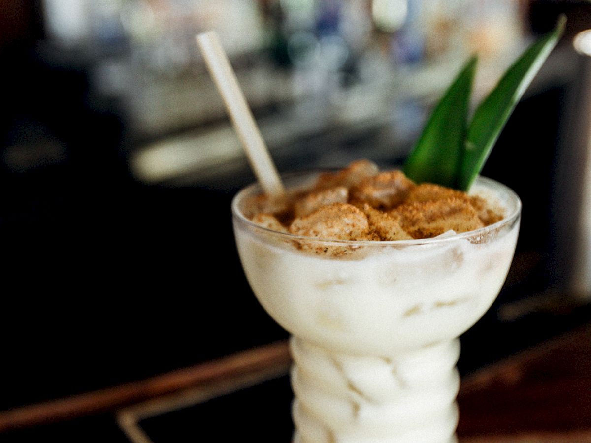 A creamy cocktail in a tall glass with cereal on top and a straw, garnished with green leaves, sits on a bar counter.
