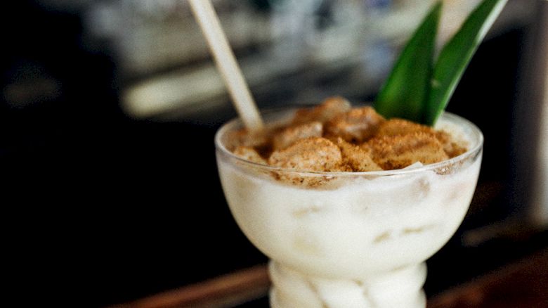A creamy cocktail in a tall glass with cereal on top and a straw, garnished with green leaves, sits on a bar counter.
