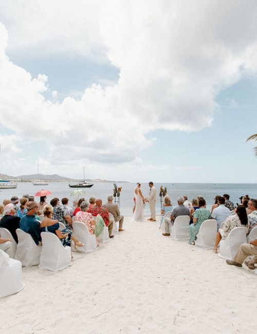 A beach wedding ceremony with guests seated on the sand, palm trees, and the ocean in the background under a partly cloudy sky.