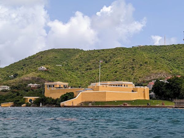 A coastal fort with yellow buildings sits by the sea against a green hillside under a blue sky with clouds.