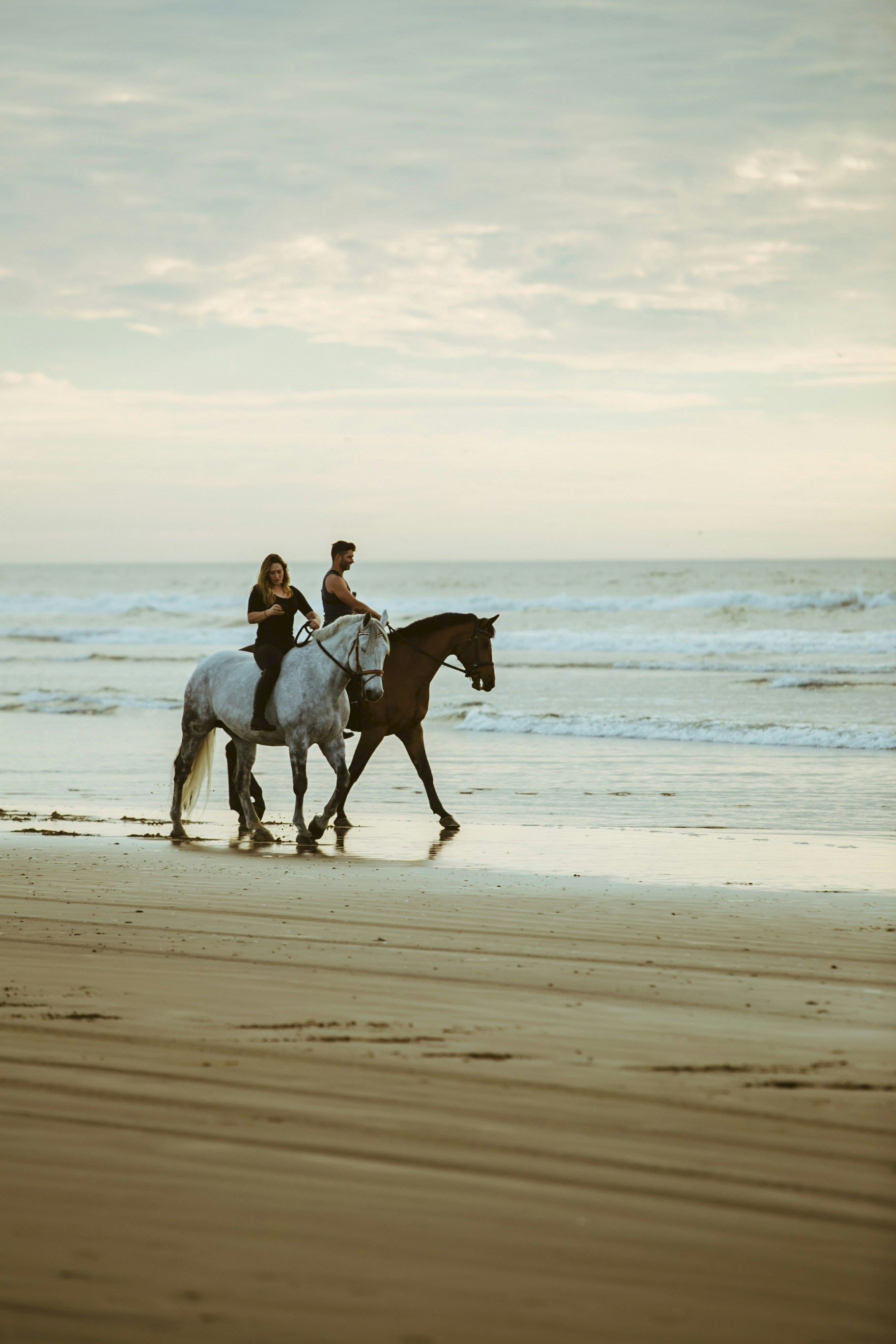 Two people riding horses along a sandy beach with waves in the background, under a cloudy sky.