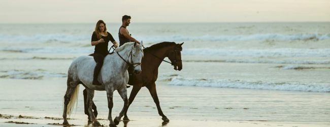 Two people ride horses along a serene beach with gentle waves under a cloudy sky, creating a peaceful and scenic atmosphere.