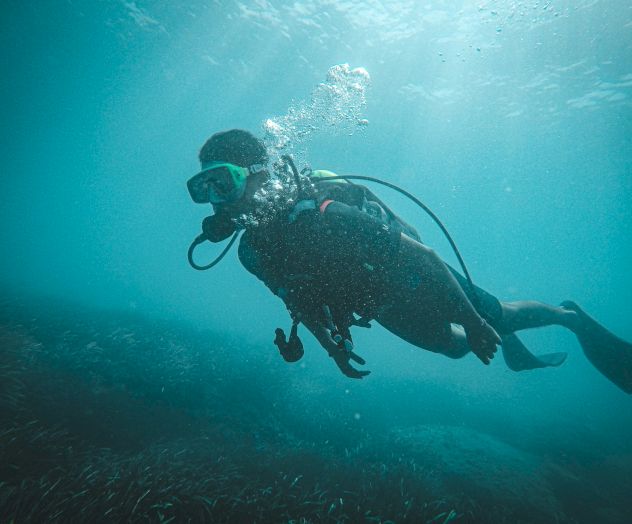 A scuba diver swims underwater in clear blue water, surrounded by bubbles, wearing a mask and fins, exploring the ocean landscape.