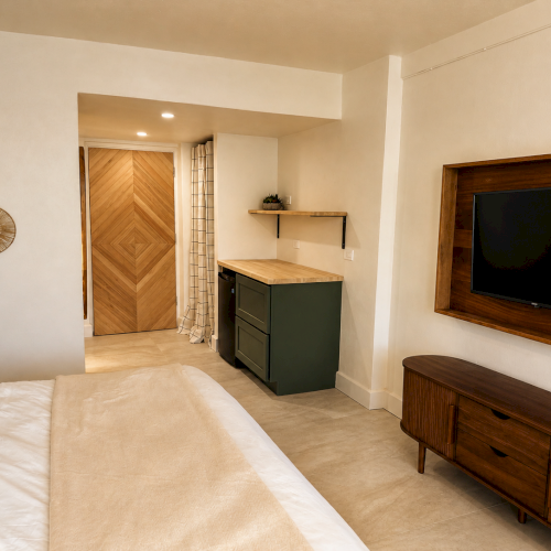 Modern bedroom with a bed, wooden TV wall, and a small built-in console/kitchenette beside a wooden door, styled cleanly.