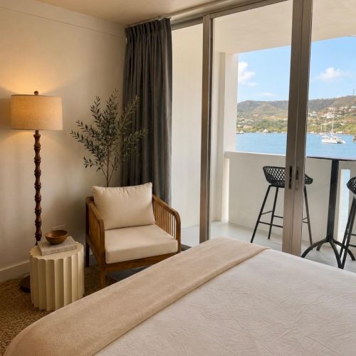Sunlit hotel room with a bed, chair, floor lamp, curtains, and a balcony view of a harbor and hills.