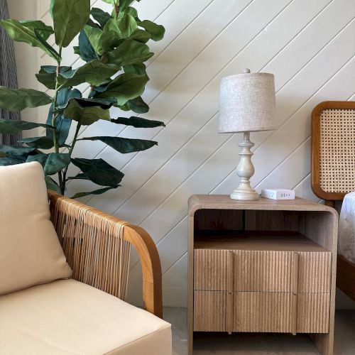 A cozy living room corner with a potted plant, beige sofa, wooden side cabinet, and a table lamp against white paneled walls.