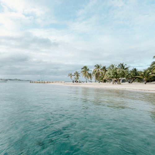 A serene beach scene with clear water, palm trees, and a few boats anchored near the shore under a blue sky with clouds.