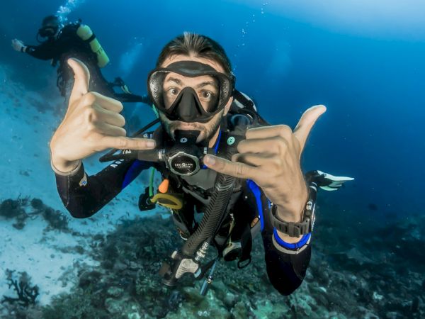 A scuba diver underwater gives a "shaka" hand sign, wearing full diving gear, with another diver visible in the background amidst ocean scenery.