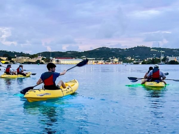 Kayakers paddle yellow kayaks across calm water toward a lit shoreline and hills under a cloudy sky.