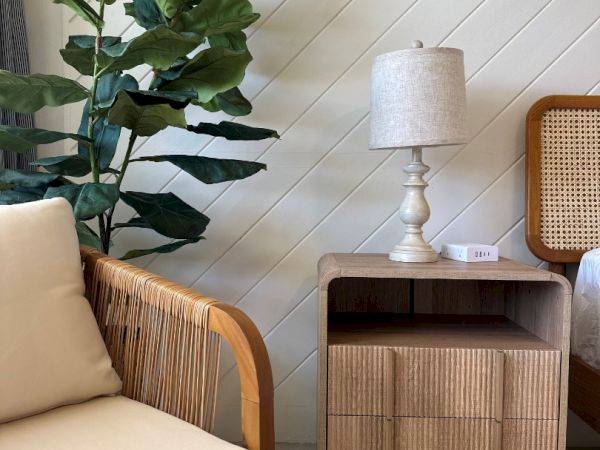 A cozy corner with a rattan armchair, tall green plant, small cabinet, and a white-shaded table lamp.