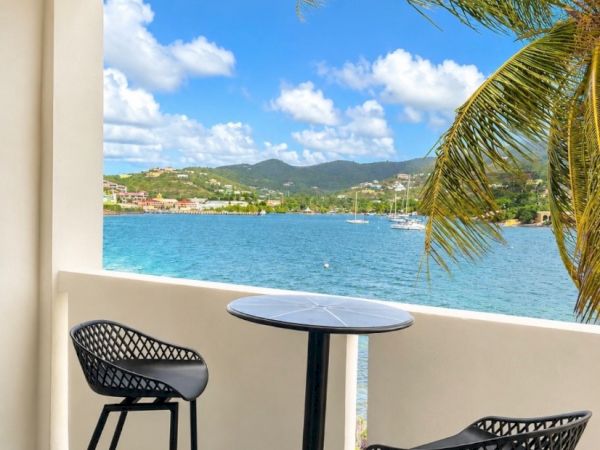 A sunny balcony with two black bar stools and a round table, looking out over the turquoise bay with palm fronds and boats.