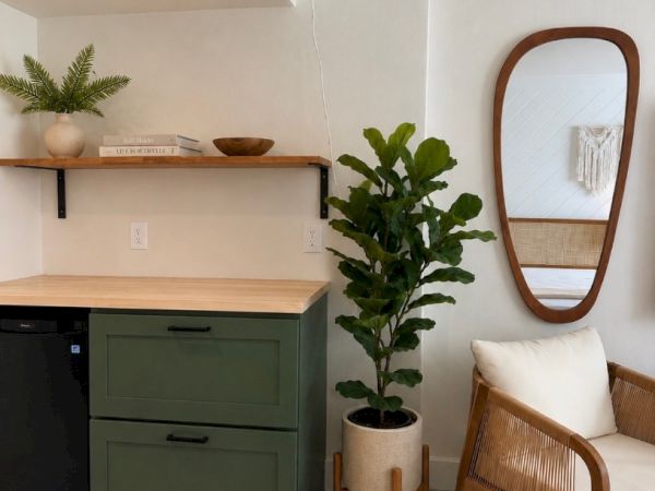 Bright modern living room: green cabinet, floating shelves, potted plant, rattan chair, and oval wall mirror.