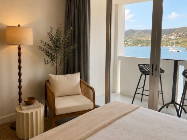 A bright hotel room with a bed, chair, floor lamp, and a balcony view of a calm bay and hills.