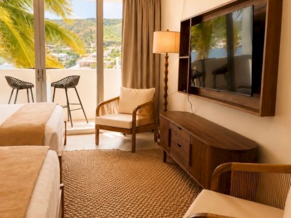 A bright hotel room with two beds, chairs, a lamp-lit TV stand, and a balcony view of hills and palm trees.
