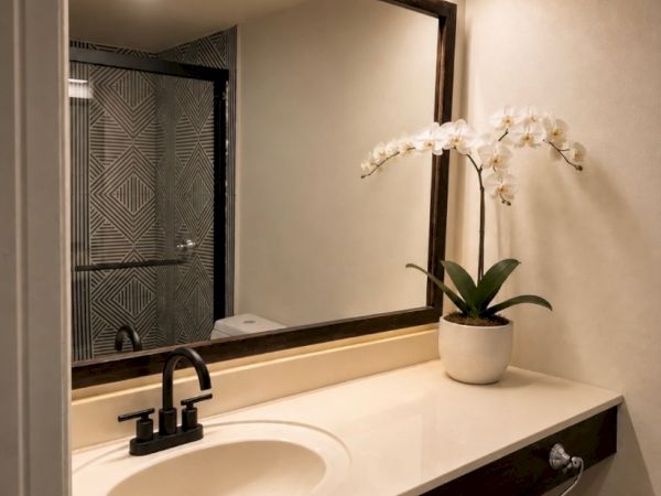 A modern bathroom vanity with a wall mirror, dual faucets, a white countertop sink, and a potted white orchid on it.
