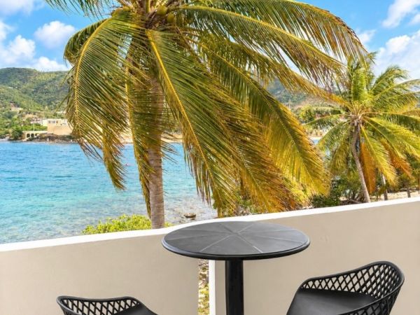 A balcony with two black chairs and a round table overlooks turquoise water, palm trees, and green hills under a blue sky.