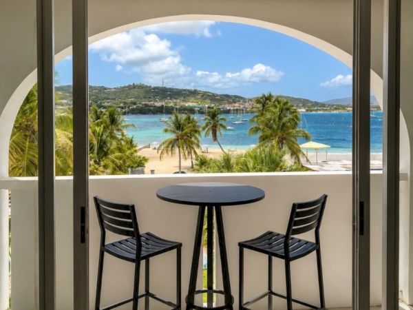 Balcony with a round table and two stools, looking out at a turquoise beach, palm trees, and boats beyond.