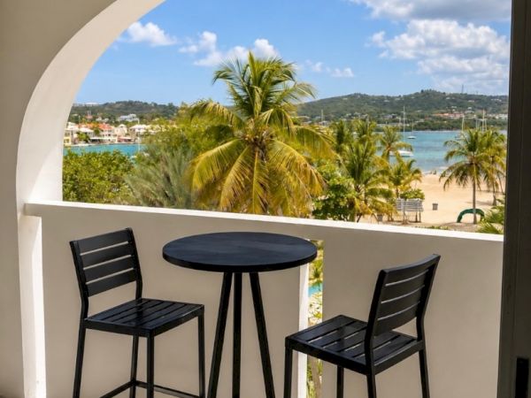 Balcony with two black stools and a round table, overlooking palm trees and a blue bay under a sunny sky.