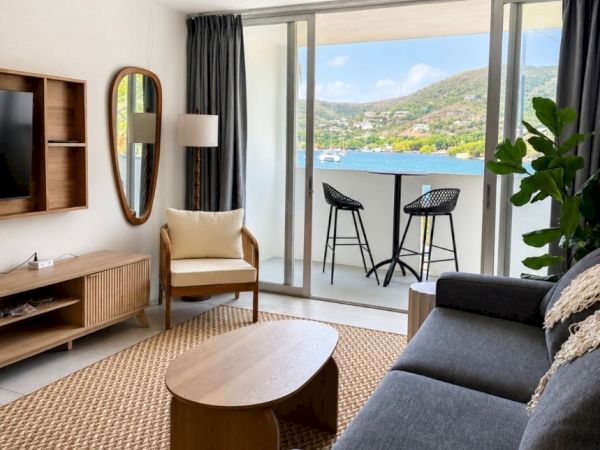 Bright living room with gray sofa and armchair, oval coffee table, ceiling fan, and large windows overlooking a lake and hills.