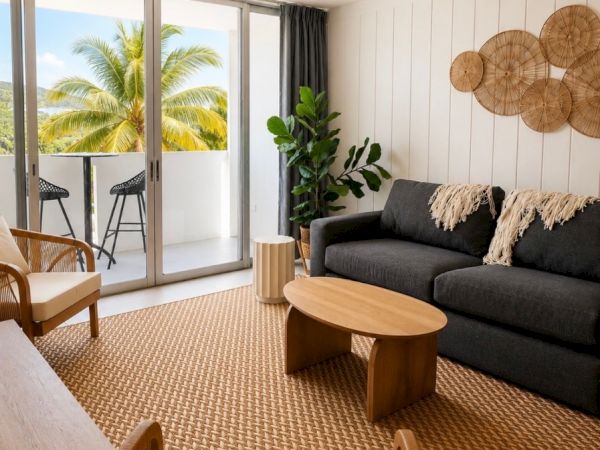Bright modern living room with rattan decor, sofa, coffee table, ceiling fan, and palm views through big glass doors.