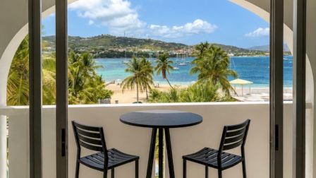 A balcony with two chairs and a round table overlooks a tropical blue sea, palm trees, and a sunny coastline.