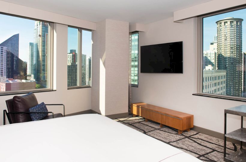 A modern hotel room with large windows offers a city view, featuring a bed, chair, wall-mounted TV, and wooden console.