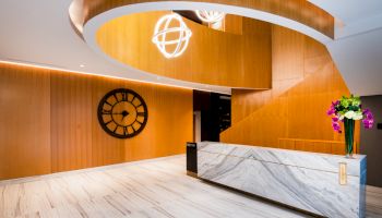A modern lobby with a curved wooden ceiling, large clock, marble reception desk, and decorative lighting and flowers.