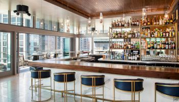 This image shows a modern bar with a selection of bottles, high stools, and large windows overlooking a cityscape.