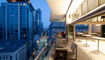 A rooftop patio with tables and chairs overlooks a city street lined with tall buildings, under a twilight sky.