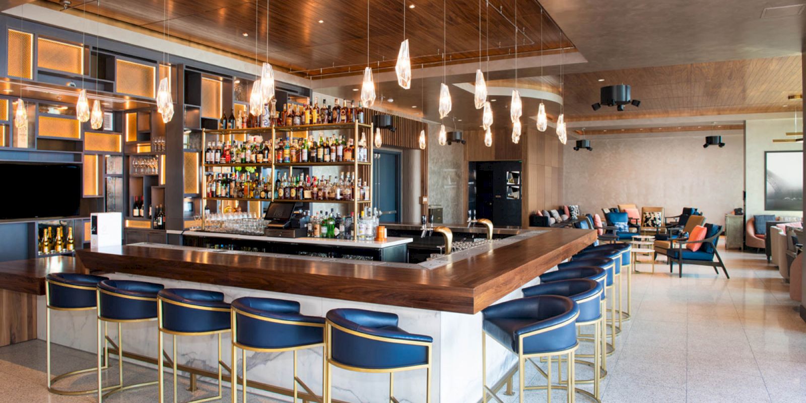 A modern bar with a wooden top, black chairs, suspended lights, and a wide selection of bottles on shelves, in a spacious setting.