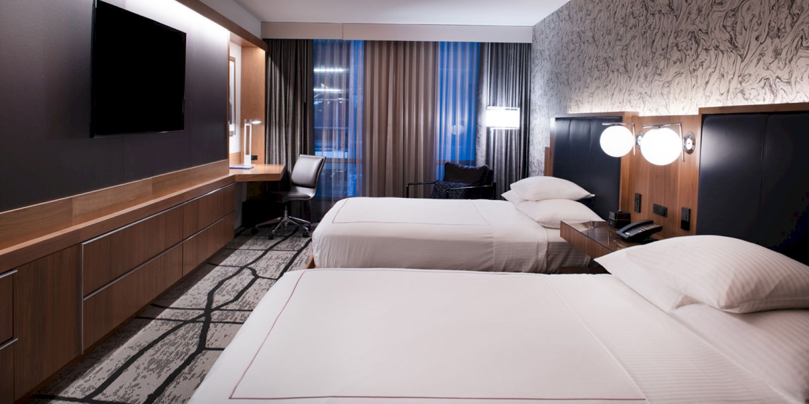 The image shows a modern hotel room with two beds, a wall-mounted TV, desk, chair, and stylish decor in a calm setting.