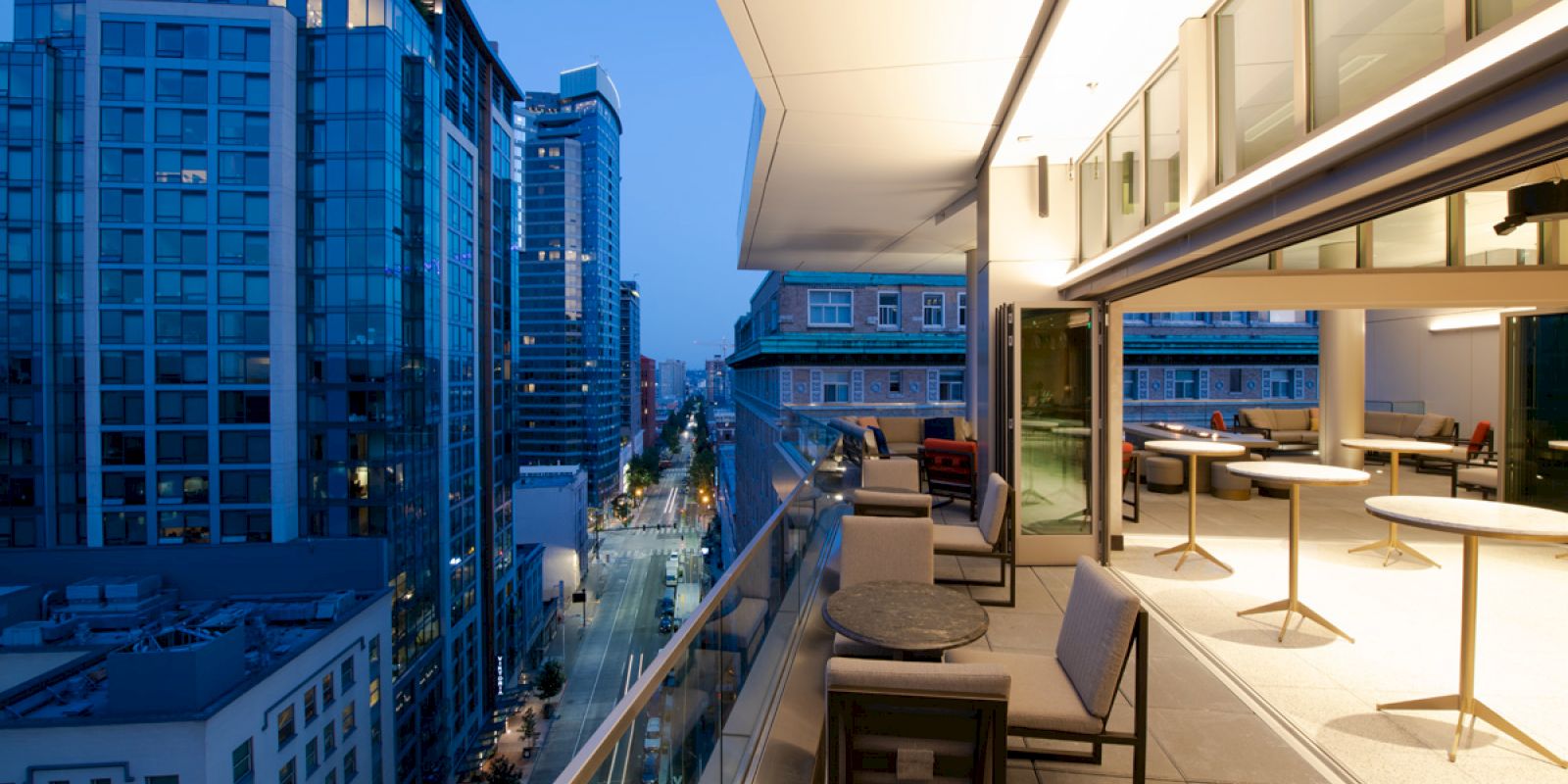 A modern cityscape view from a balcony at twilight, featuring high-rise buildings and an outdoor seating area with tables and chairs.