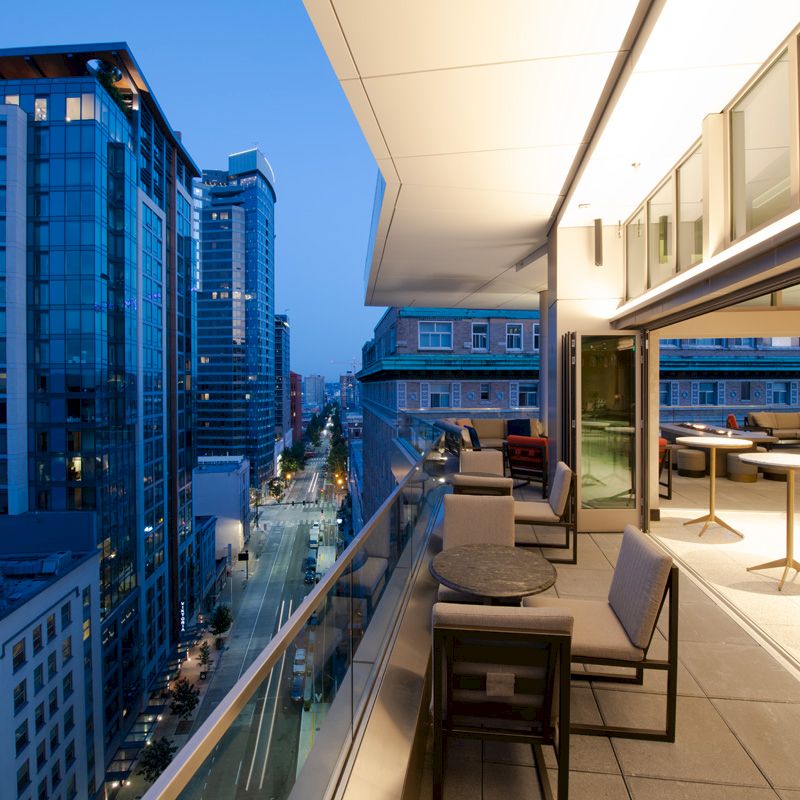 A modern cityscape at dusk with tall buildings and a balcony featuring tables and chairs.