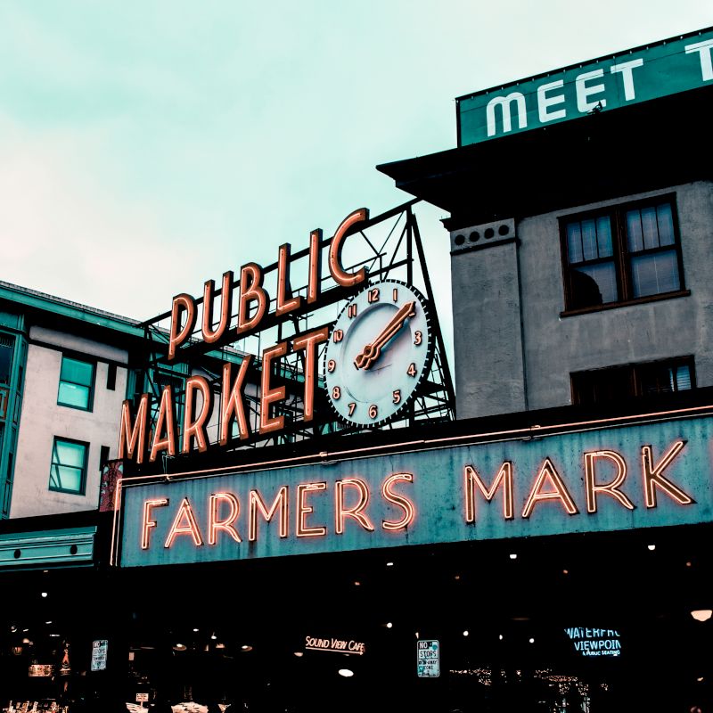 This image shows a neon sign for a public and farmers market, with a clock and surrounding buildings, under a cloudy sky.