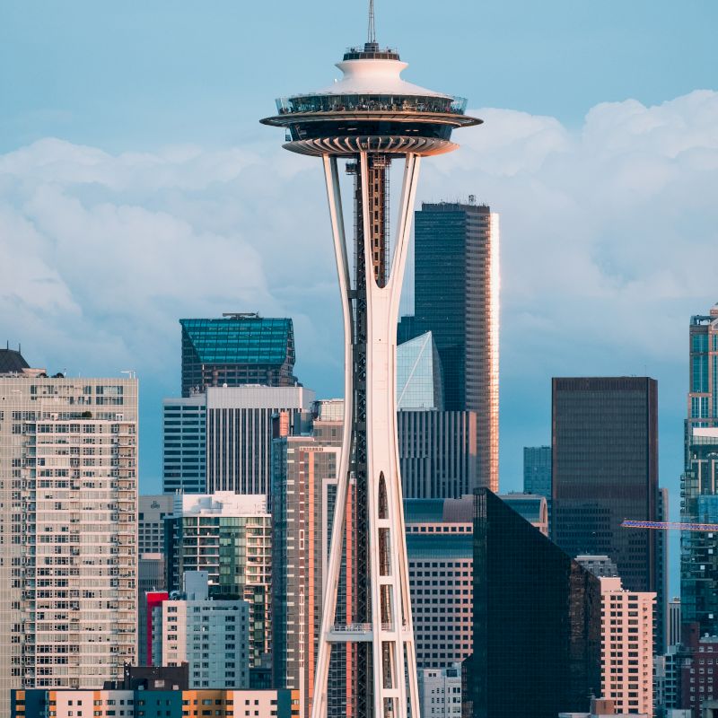 The image shows the Space Needle, a prominent observation tower, with a backdrop of modern buildings and a cloudy sky.
