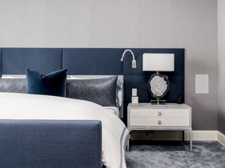A modern bedroom with a blue and white color scheme, featuring a bed, pillows, nightstand, lamp, and decorative accents.