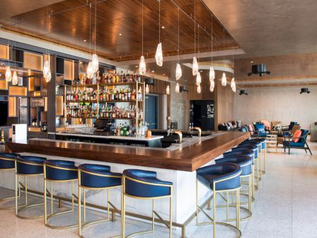 The image shows a modern bar with blue chairs, a hanging light fixture, and a well-stocked liquor shelf, set in a stylish lounge area.
