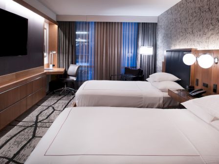 The image shows a modern hotel room with two beds, a TV, a desk, and contemporary decor, featuring a patterned accent wall and carpet.