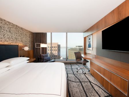 A modern hotel room with a large bed, desk, and TV features a view of buildings and a body of water through a wide window.