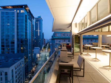 A modern balcony overlooking a cityscape at dusk, featuring seating areas and tables, illuminated by warm indoor lighting.