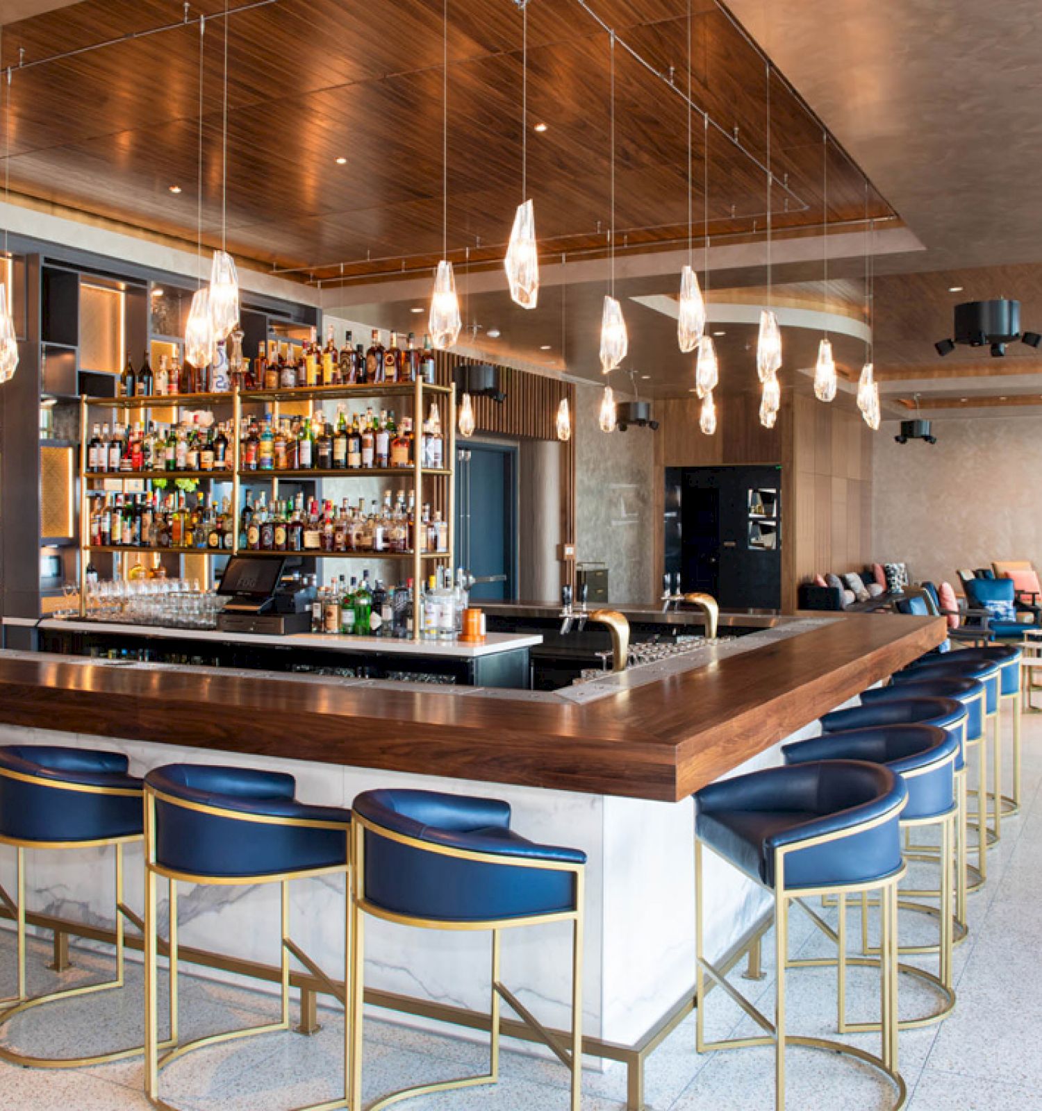 A modern bar with a wooden counter, blue stools, hanging lights, and shelves of liquor bottles. There's seating in the background.
