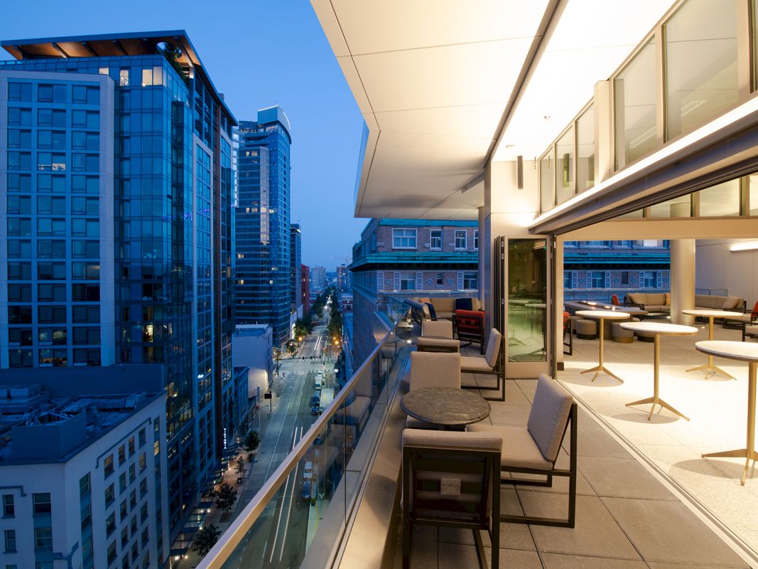 A modern balcony view overlooking an urban street and tall buildings at dusk, featuring outdoor seating and warm interior lighting.