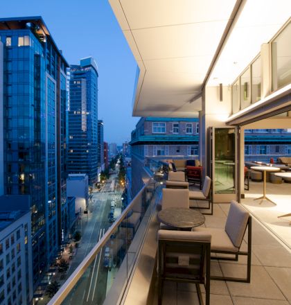 A modern cityscape with tall buildings is viewed from a balcony with seating and tables, illuminated by evening lights.