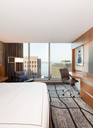 A modern hotel room features a large bed, flat-screen TV, desk with a chair, and a window view of buildings and water.