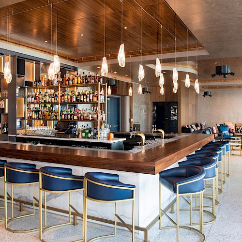 The image shows a modern bar with sleek blue chairs, a well-stocked liquor display, pendant lights, and comfortable seating in the background.