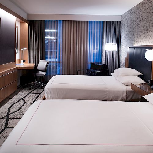 The image shows a modern hotel room with two beds, a TV, and a desk by the window with patterned curtains and a stylish carpet.