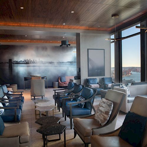 A modern lounge with plush seating, tables, and a large window view of water and a Ferris wheel under clear skies, has ambient lighting.