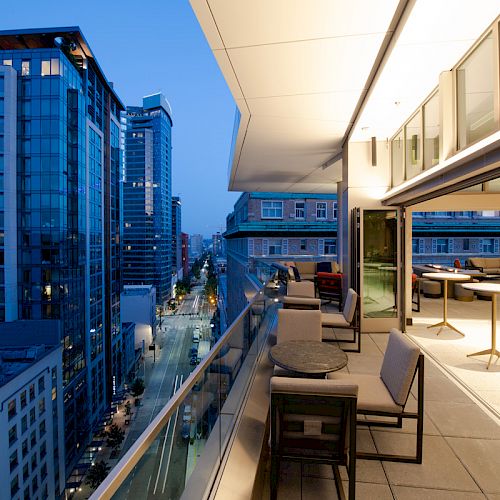 A modern urban balcony overlooks a cityscape, featuring outdoor seating and illuminated interiors at dusk.