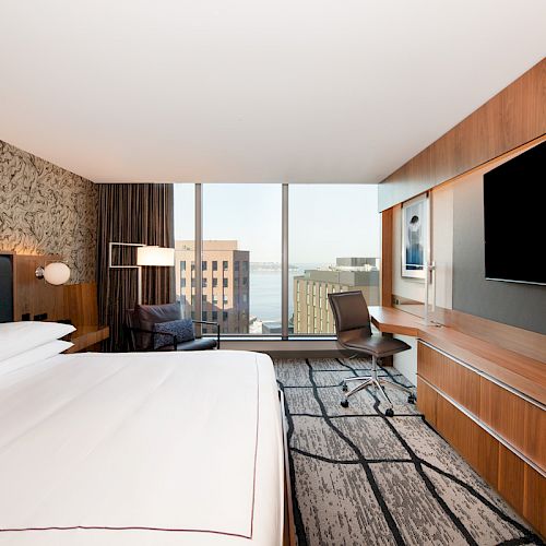A modern hotel room with a large bed, desk, chair, and flat-screen TV. The window offers a view of the city and water beyond.